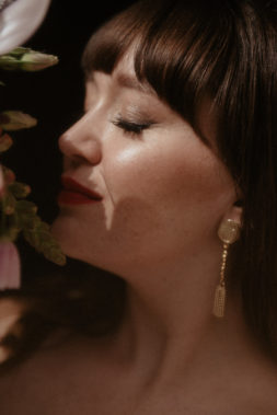 Le portrait d'une mariée de profil. Elle sent son bouquet. C'est un maquillage très lumineux et glow. Une bouche rouge et des yeux travaillés en liner fondu au marron froid et légères paillettes.