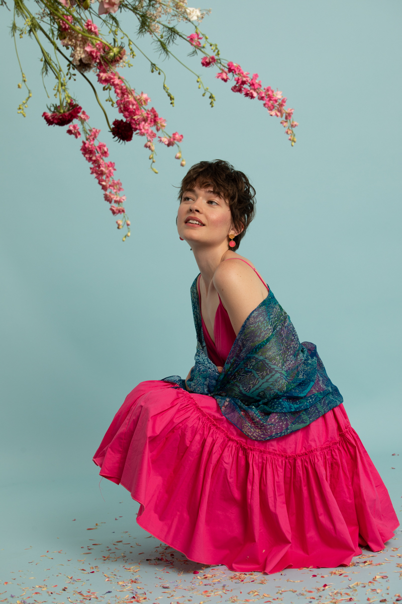 Une jeune femme chatain aux cheveux courts. Elle porte une robe rose et un foulard Petrusse sur ses épaule. Elle a un maquillage très lumineux et pop aux couleurs roses.